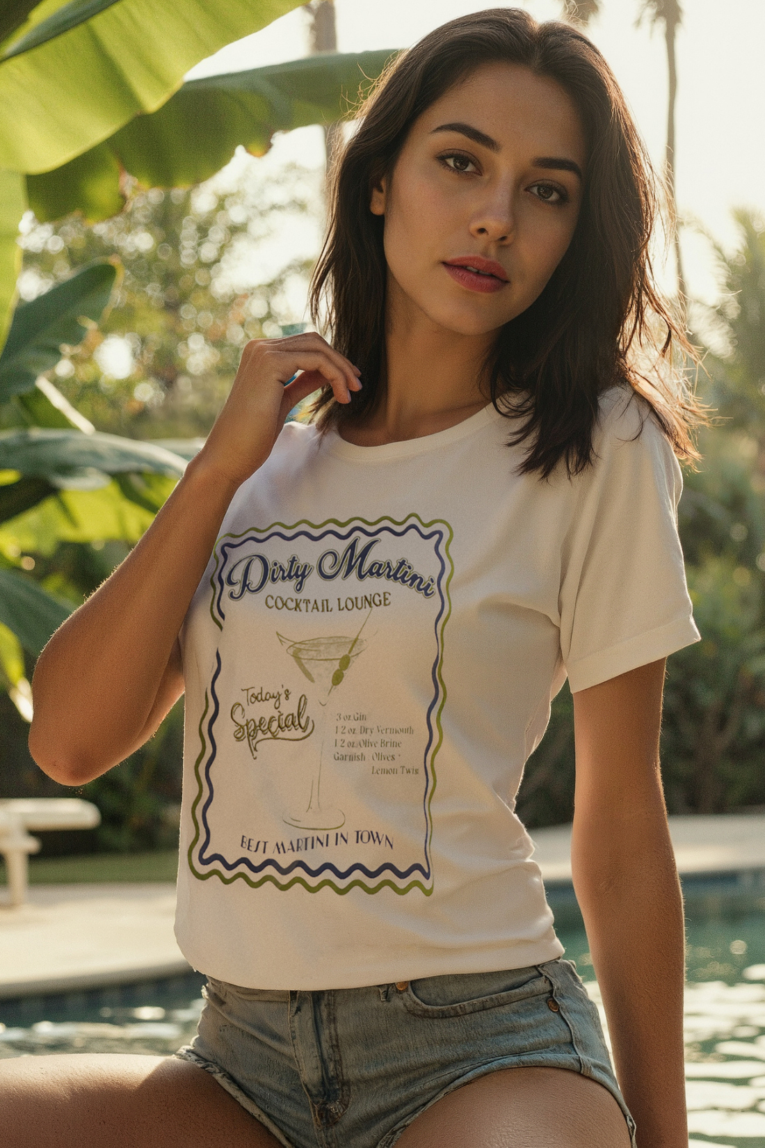 A woman wearing a white crop tee with a graphic that includes text and a martini illustration, paired with denim shorts, standing near a pool.