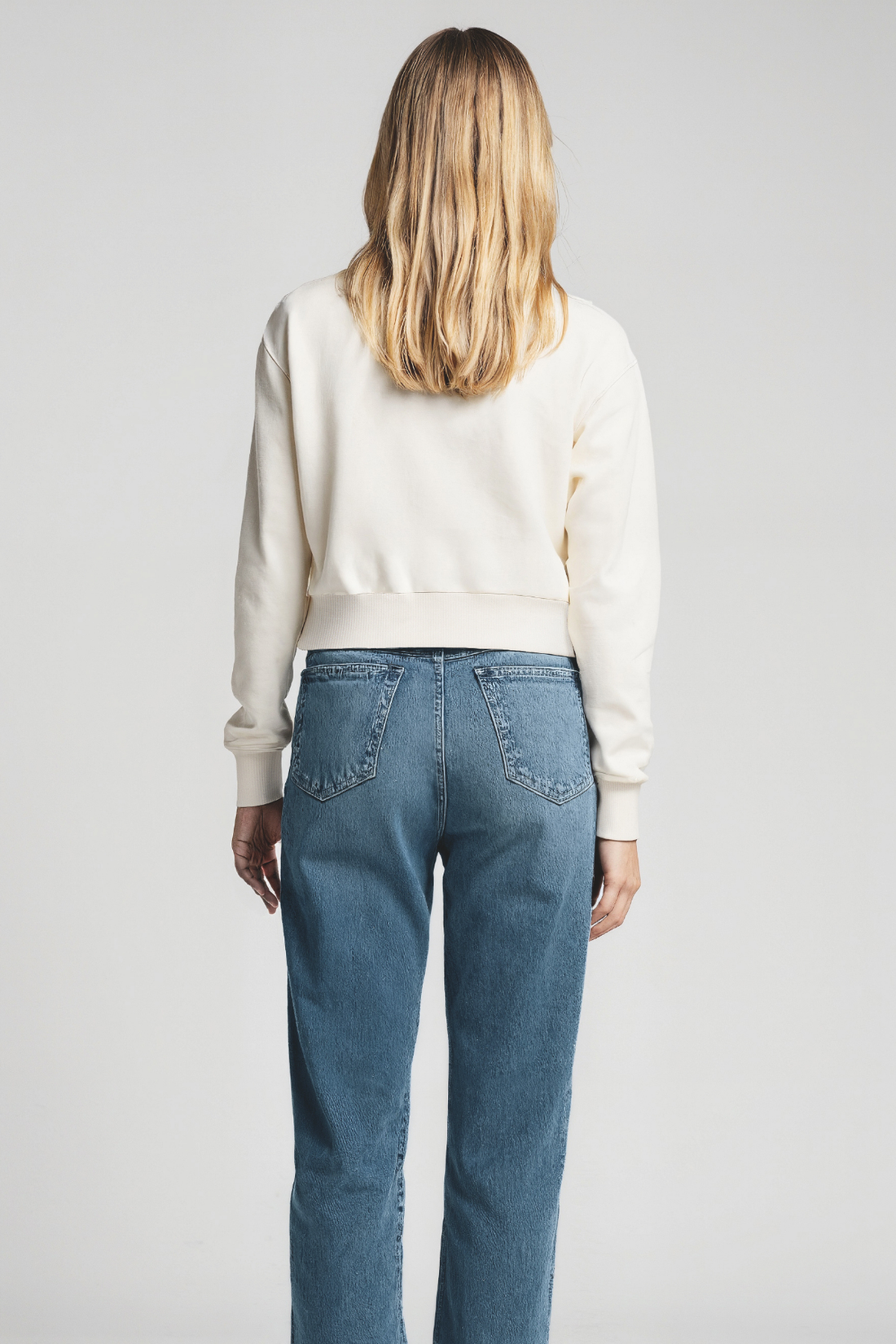 Back of women wearing a white sweater and blue jeans on a plain background