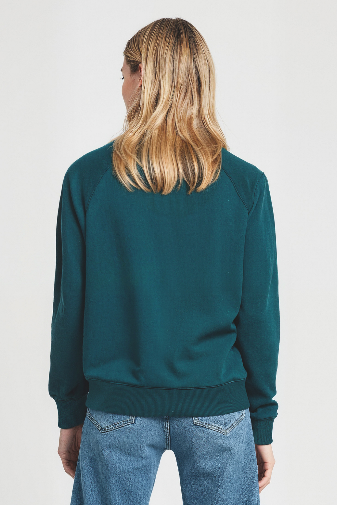 Back view of a woman wearing a teal sweatshirt and blue jeans on a white background