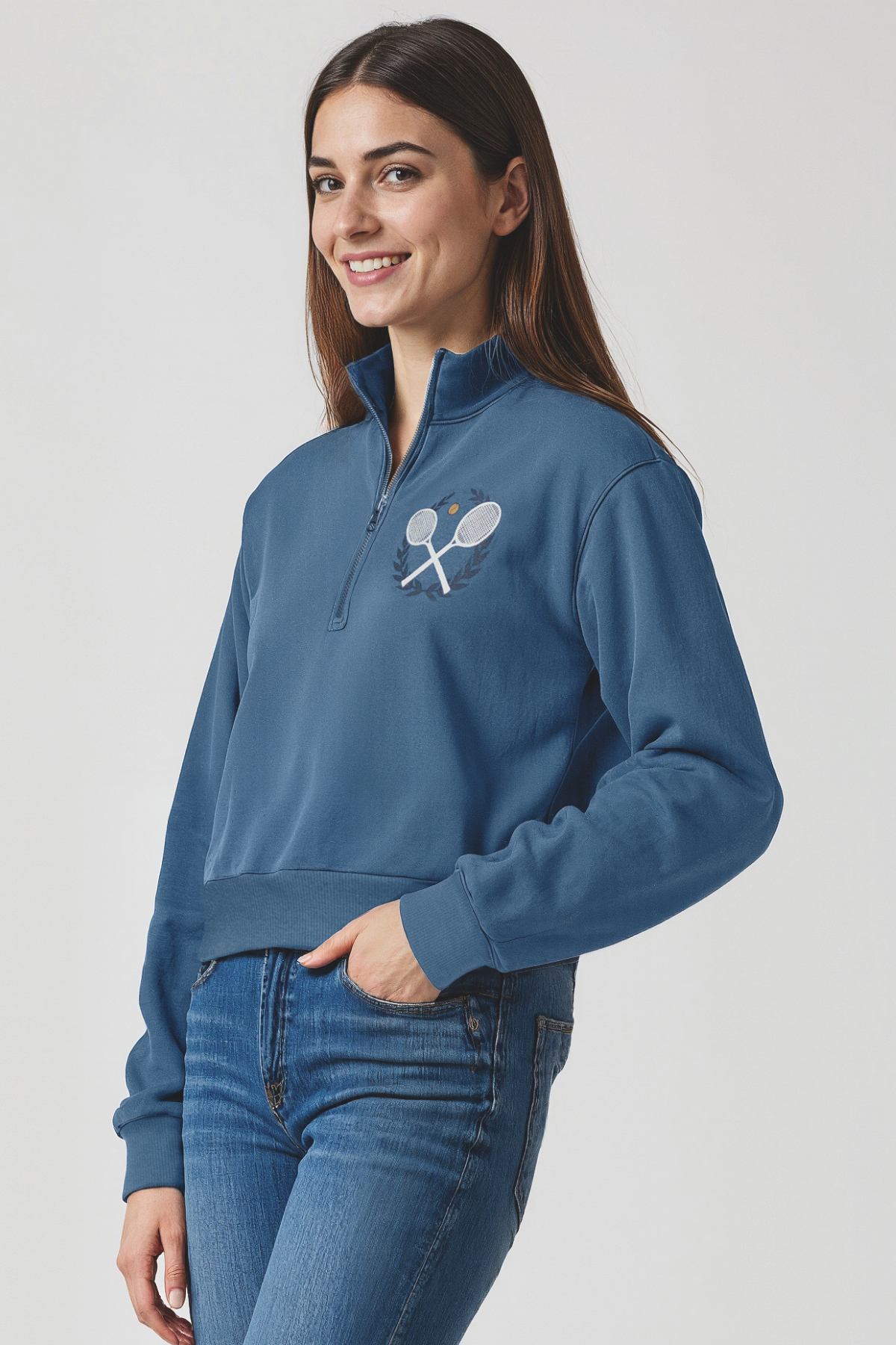 Woman wearing a blue 1/2 zip pullover with 2 tennis rackets logo on a plain background