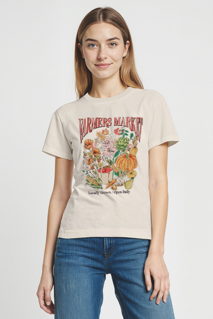 Woman wearing a beige t-shirt with a colorful graphic design of &quot;FARMERS MARKET; Locally Grown / Open Daily&quot; text on a plain background