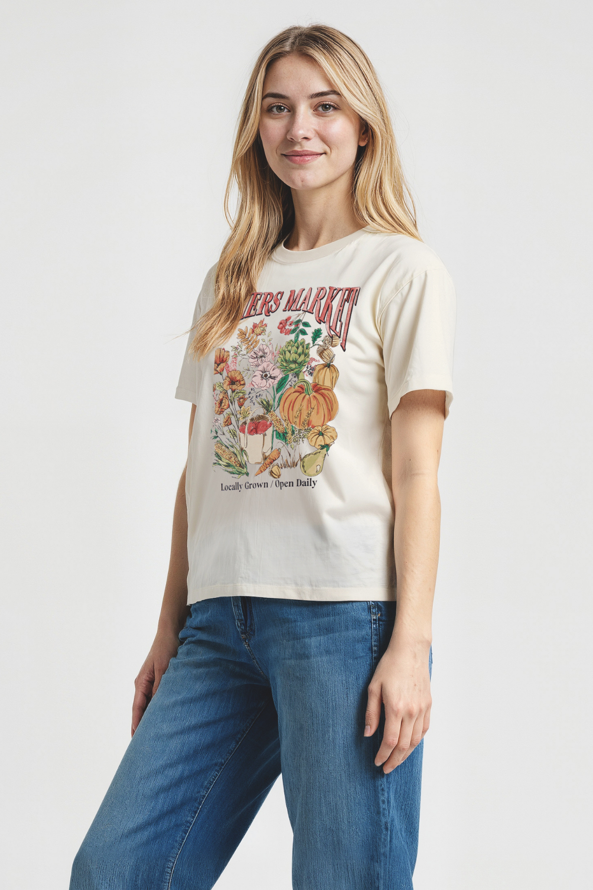 Woman wearing a beige t-shirt with a colorful graphic design of &quot;FARMERS MARKET; Locally Grown / Open Daily&quot; text on a plain background