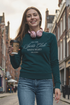 Woman holding a coffee cup and wearing headphones and a pine green sweatshirt, standing on a street.