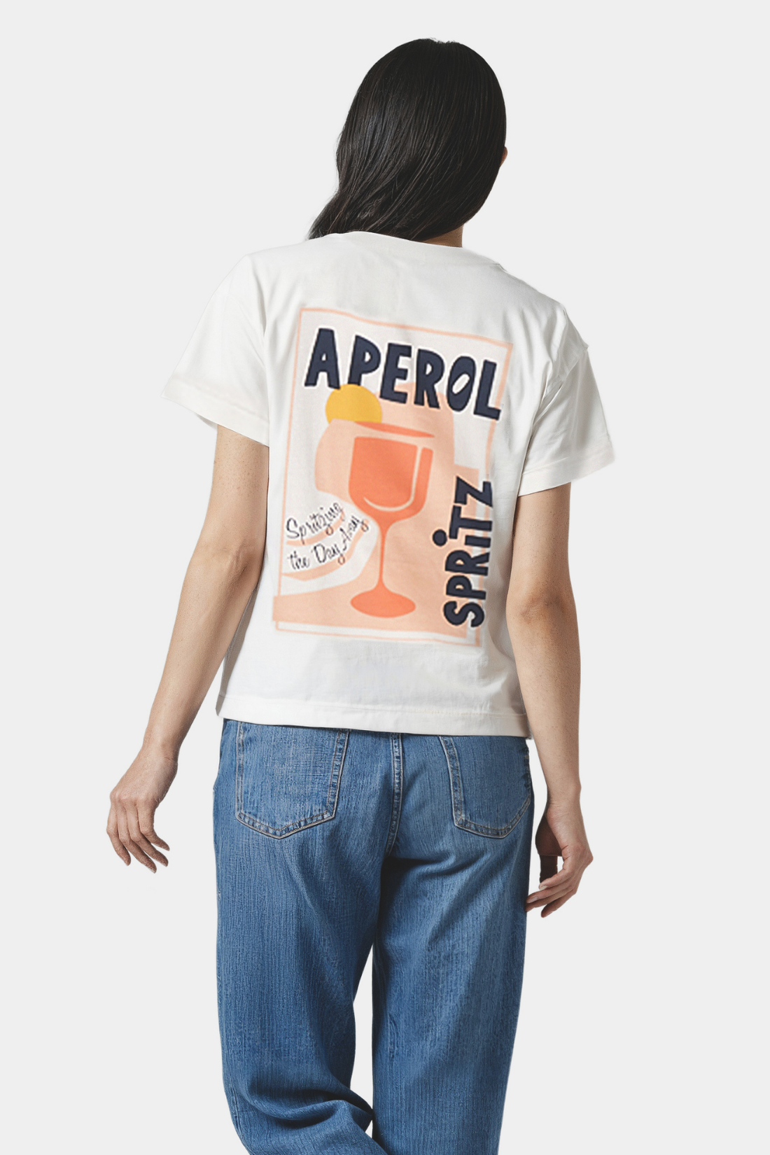 Back of woman wearing a white t-shirt with Aperol Spritz graphic on the back, standing against a plain background.