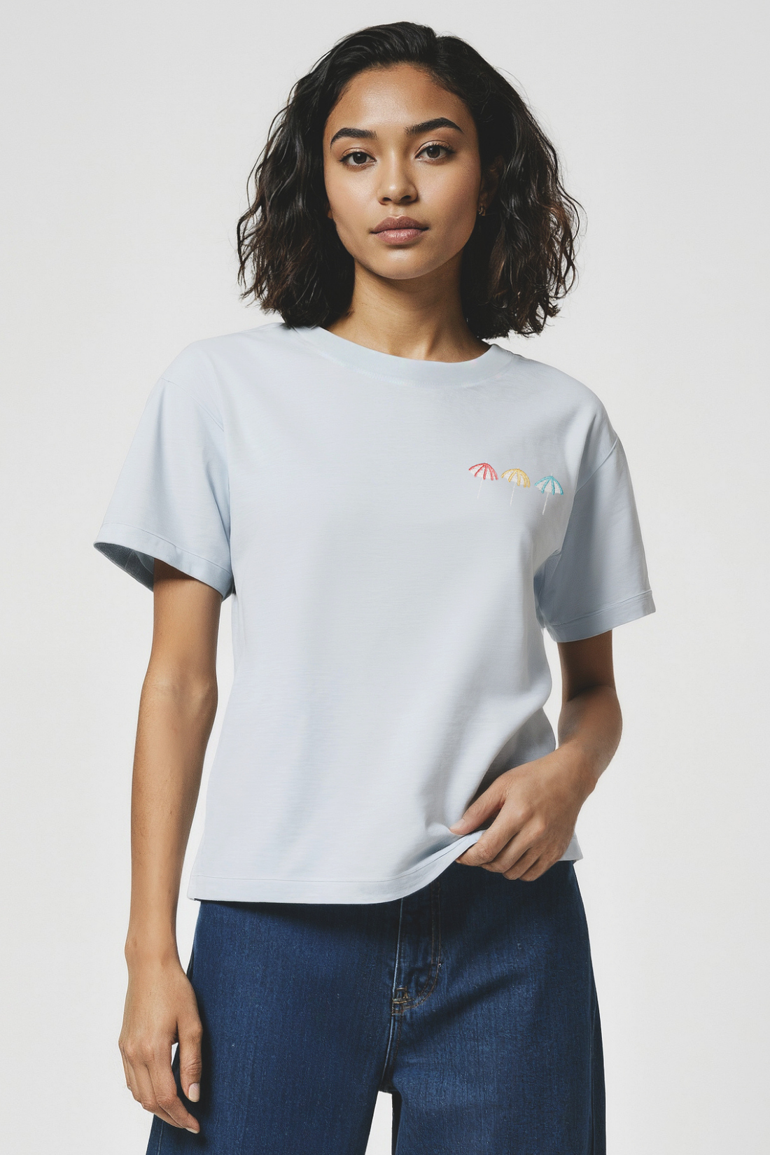 Woman wearing a light gray t-shirt with small 3 colorful embroidered beach umbrellas designs on a plain background