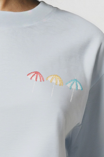 Close up of light blue shirt with 3 colorful embroidered beach umbrellas designs on a plain background