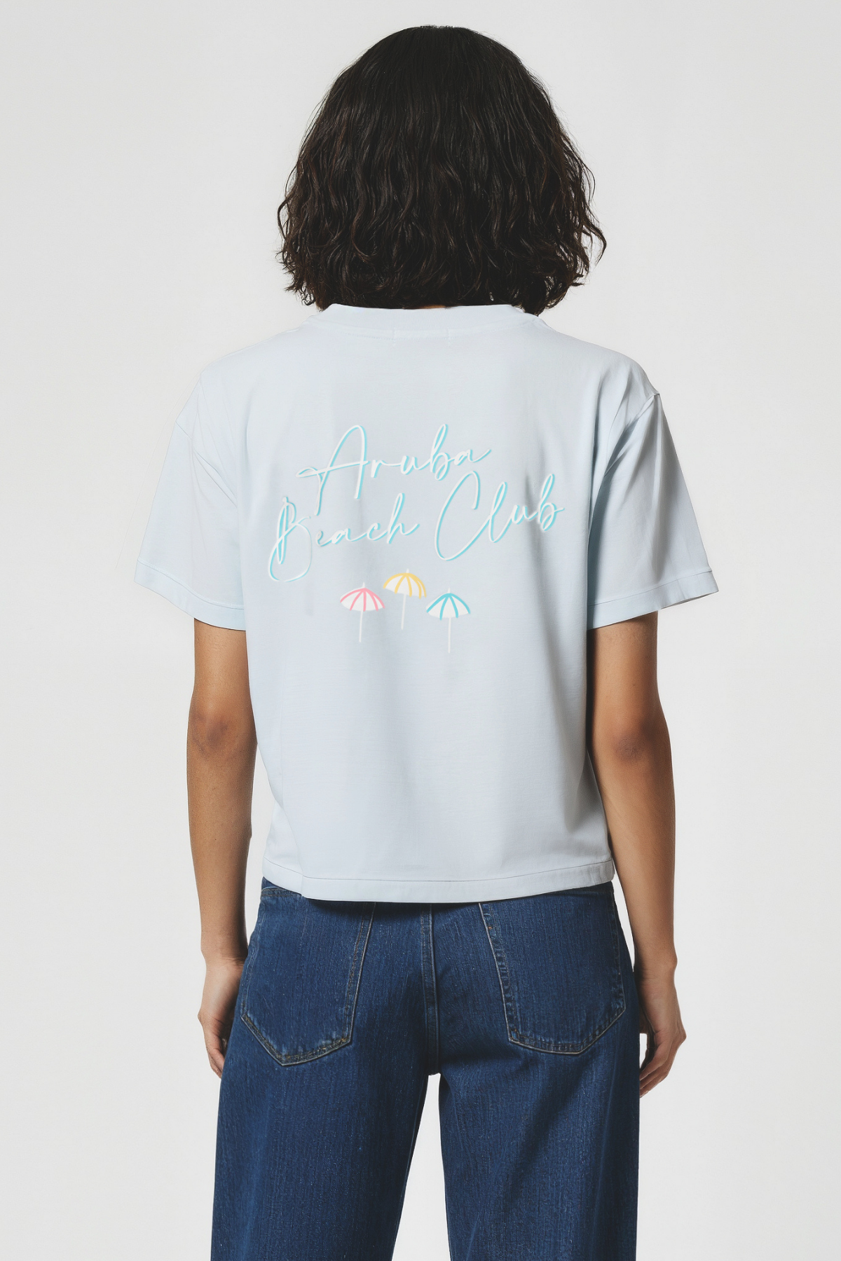 Woman wearing a light blue t-shirt with colorful text and graphics on the back, standing against a plain background.