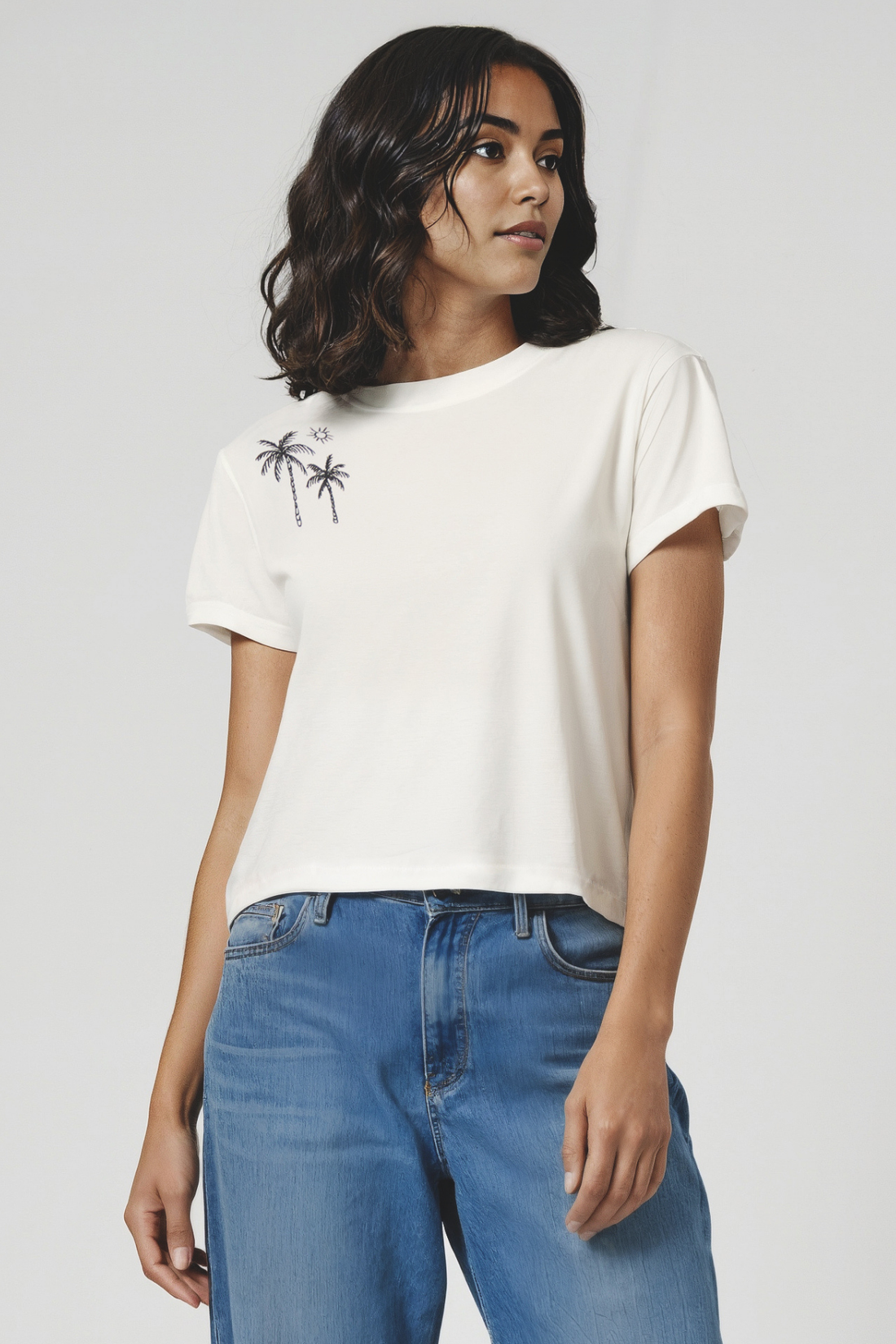 Woman wearing a white t-shirt with a palm tree design on top right shoulder and blue jeans on a plain background