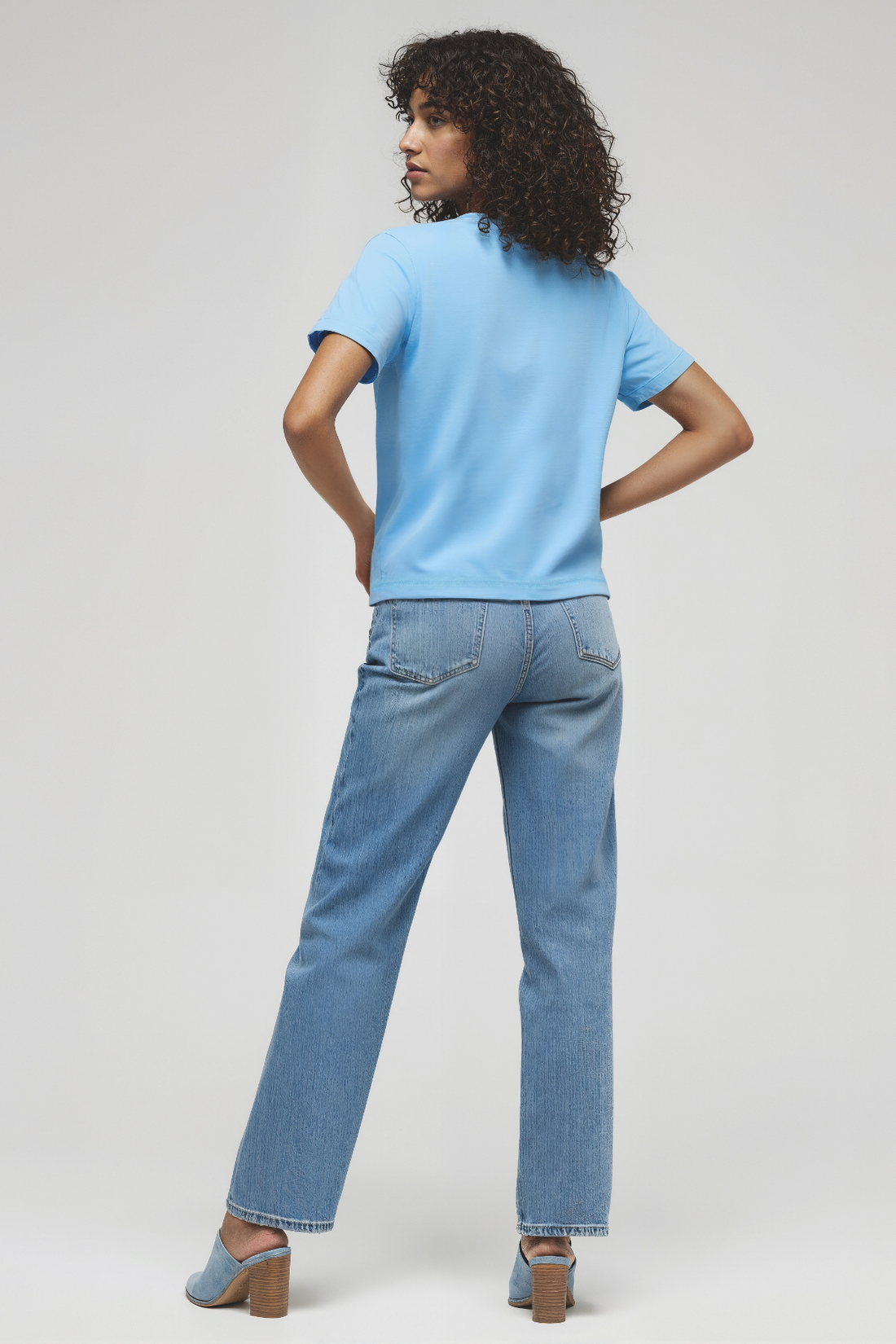 Back view of woman wearing a light blue shirt and jeans on a white background