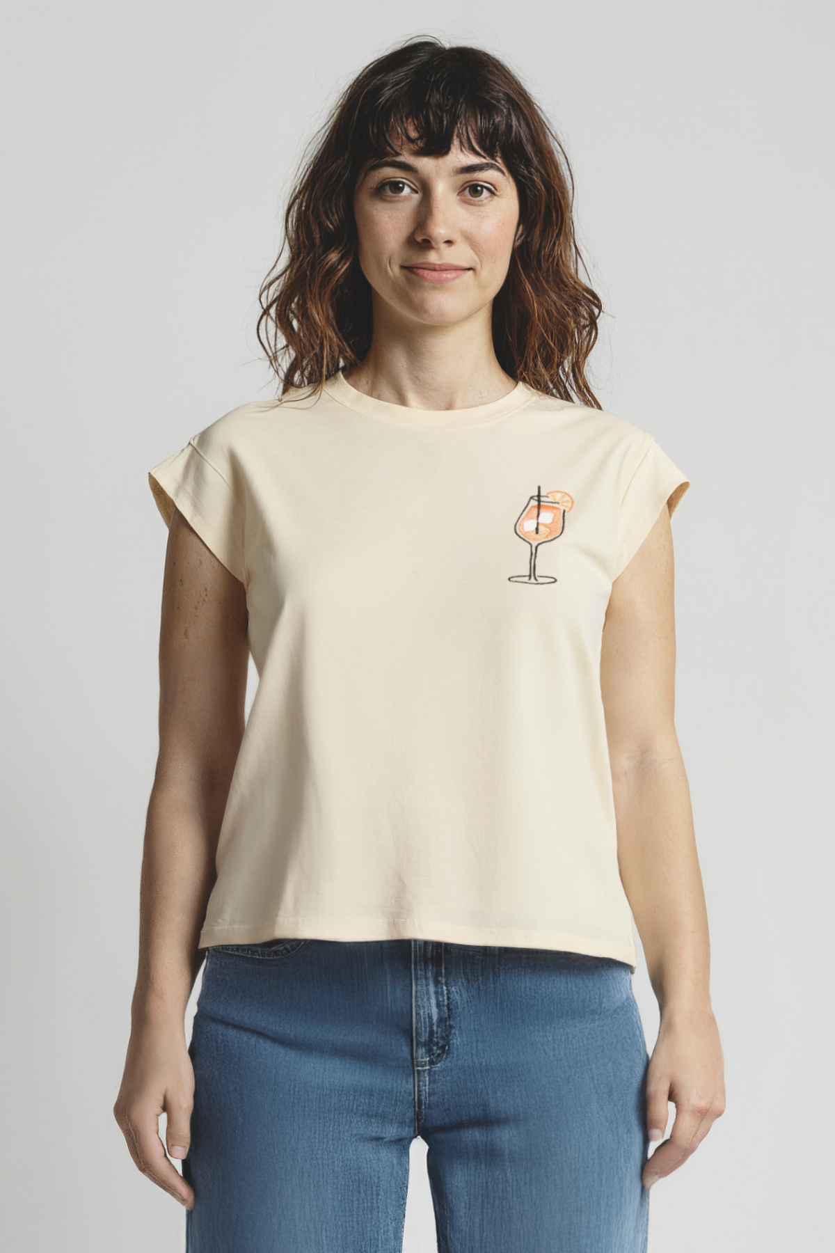 Woman wearing a yellow tank with a small cocktail drink glass graphic and jeans on a plain background
