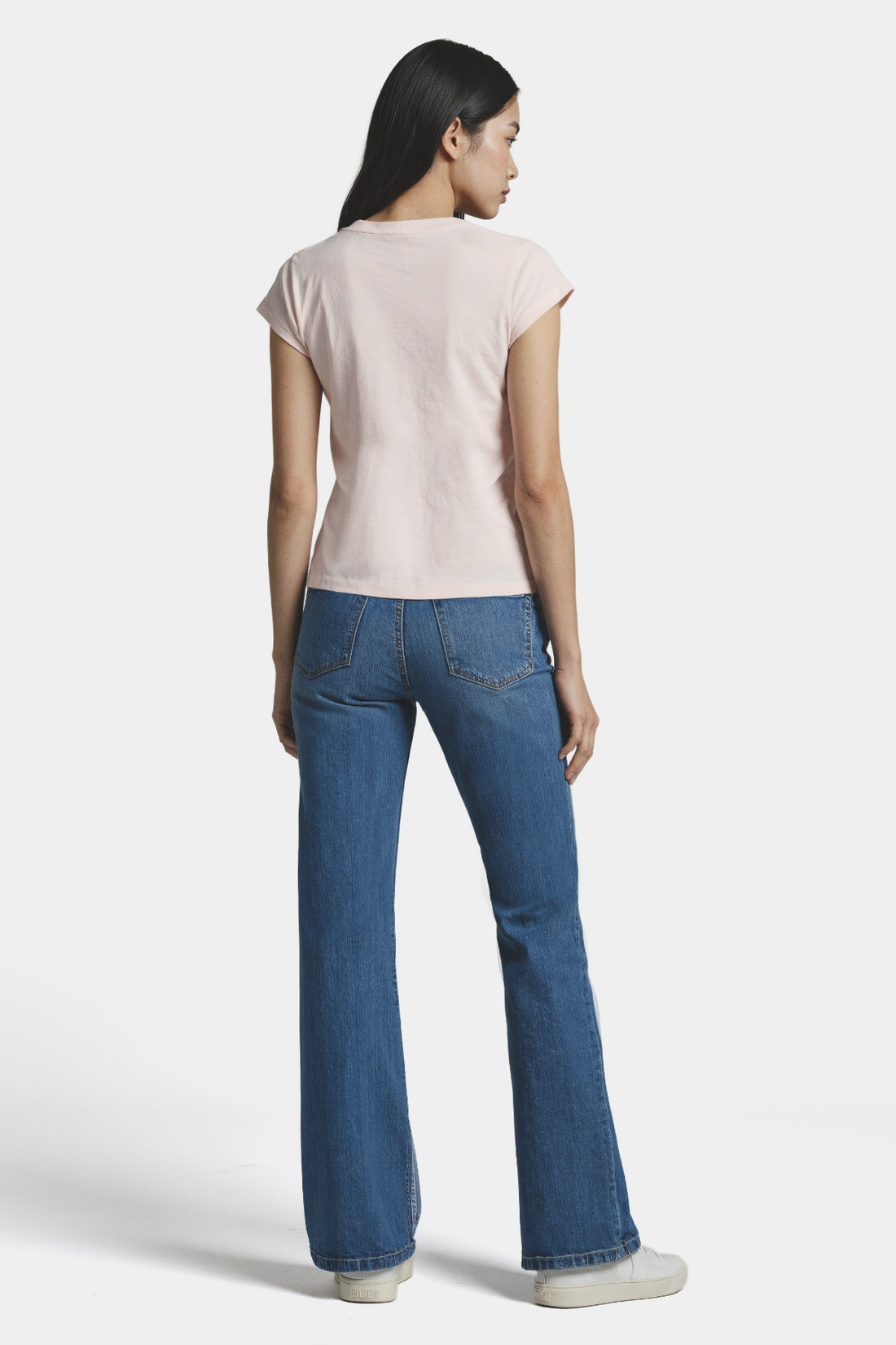 Back of woman wearing a light pink t-shirt and blue jeans on a white background.