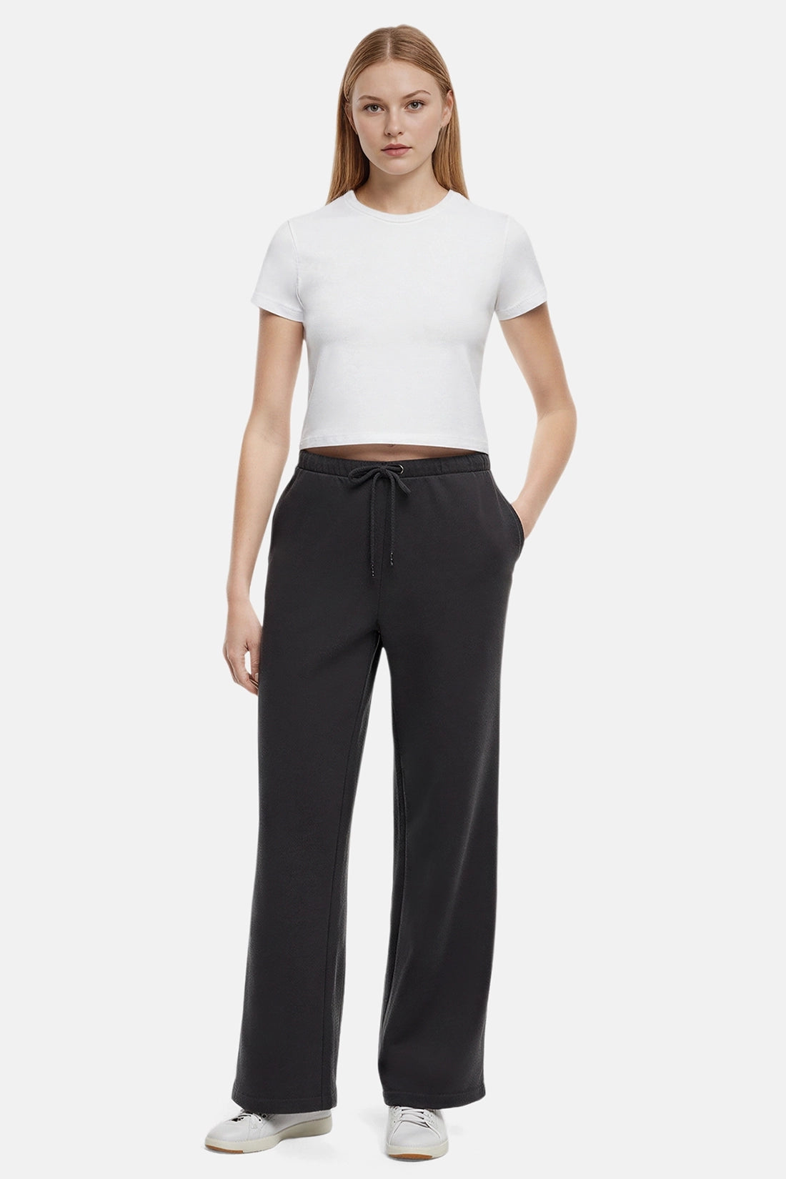 Woman wearing a white cropped t-shirt and heather black wide-leg drawstring sweatpants with hand in pocket on a white background