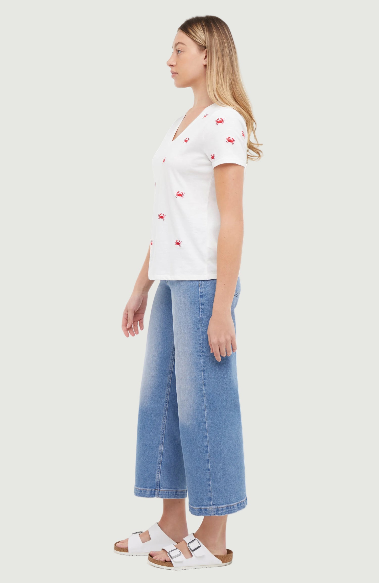 Side view of woman wearing a white t-shirt with red embroidered crab patterns and blue jeans on a light gray background