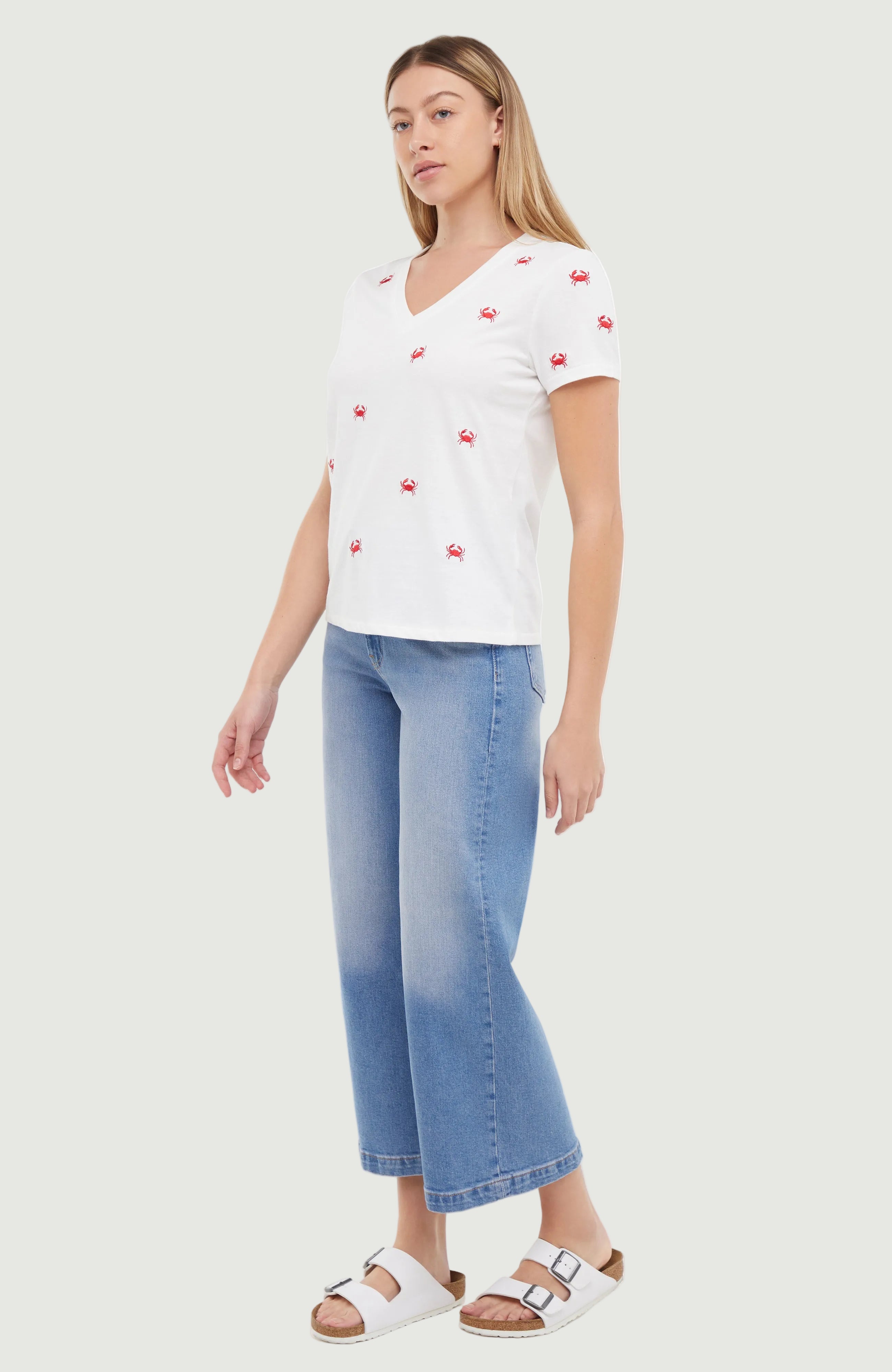 Side view of woman wearing a white t-shirt with red embroidered crab patterns and blue jeans on a light gray background