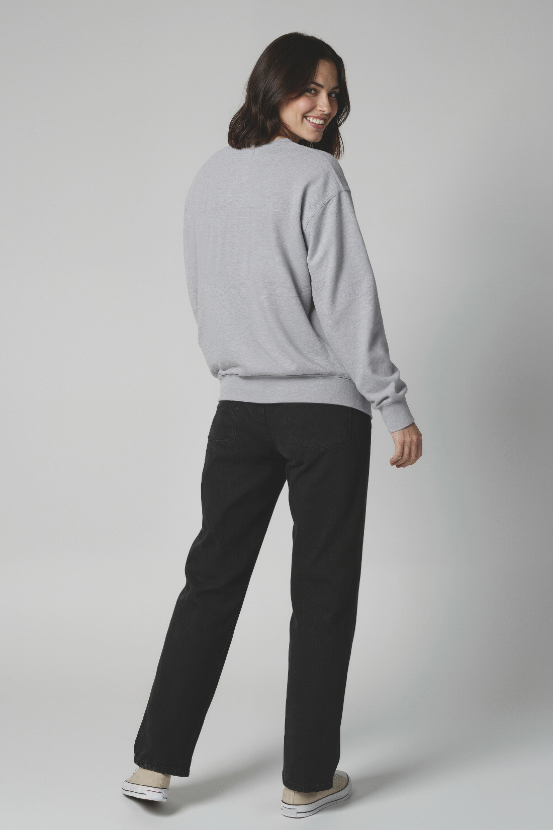 Back of woman wearing a gray sweatshirt with black jeans on a plain background