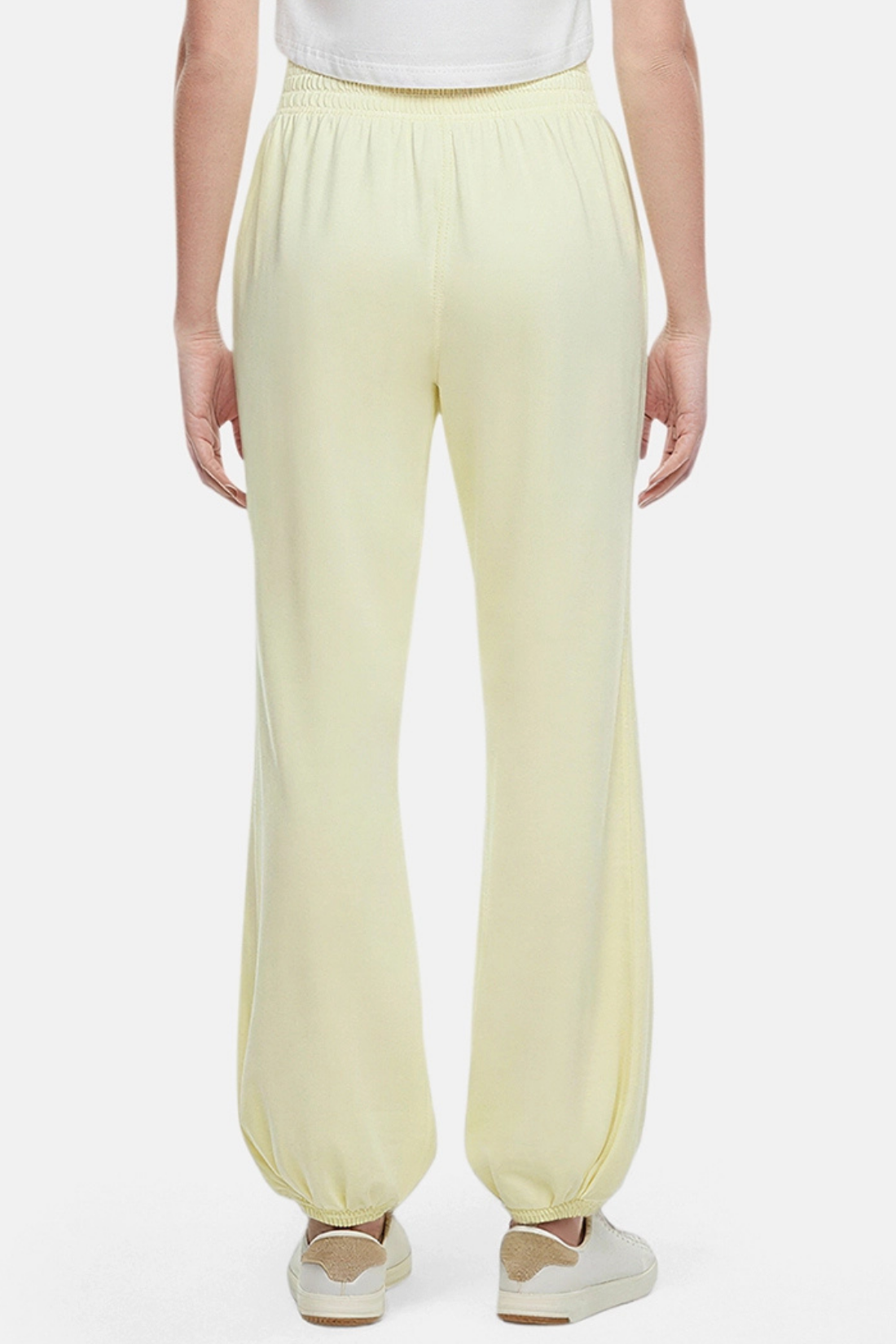 Back view of woman wearing yellow pants, white tee and white sneakers on a white background. 