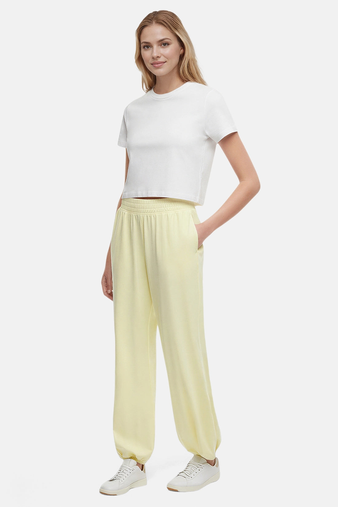Front view of woman wearing yellow pants, white tee and white sneakers on a white background. 