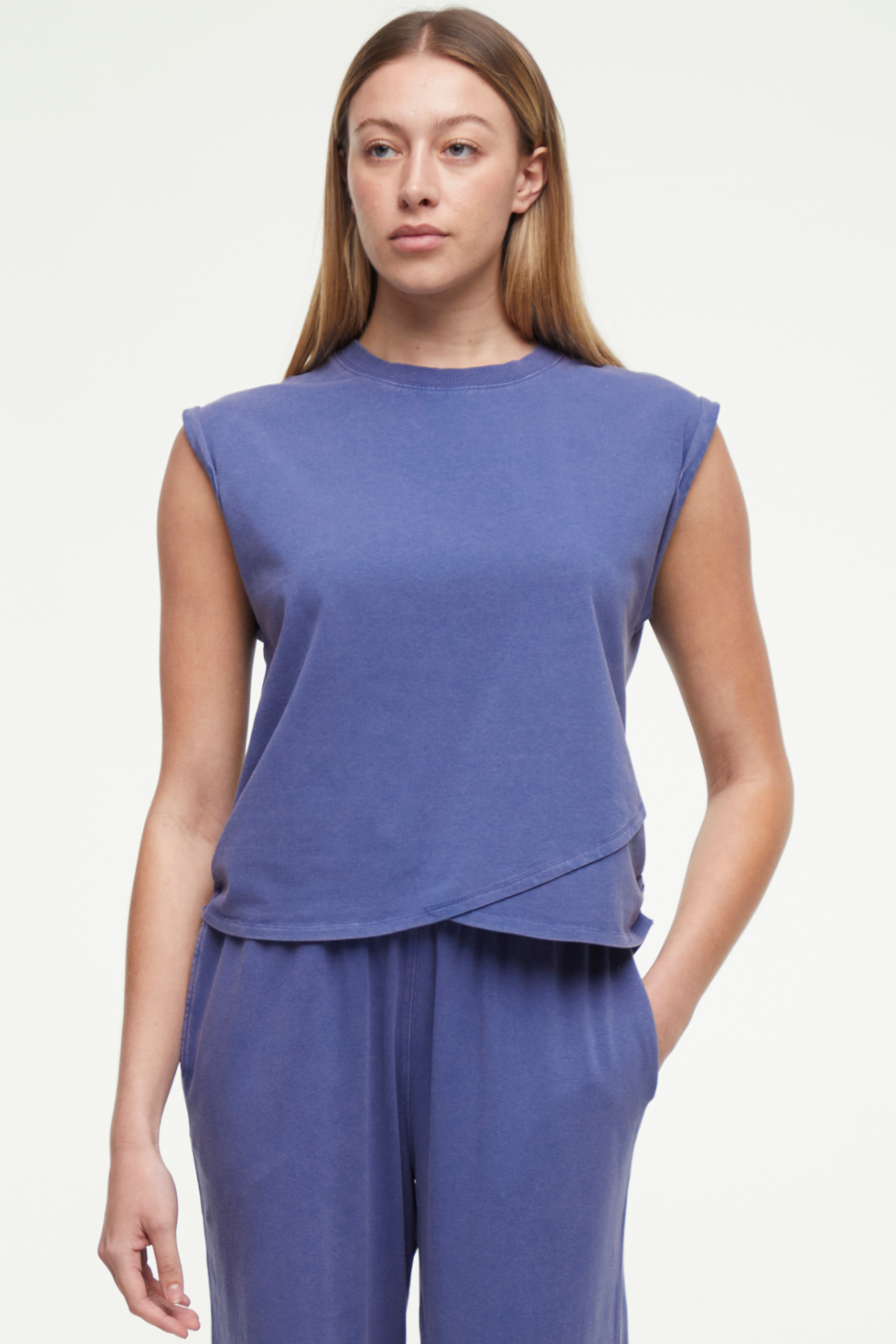 Front view of woman wearing a gray-blue sleeveless asymmetrical top and pants on a white background. 