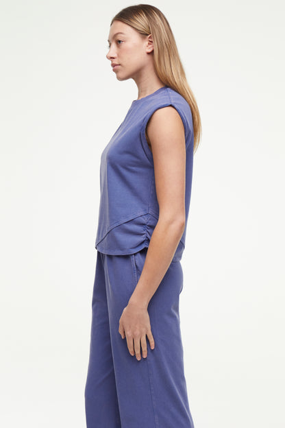 Side view of woman wearing a gray-blue sleeveless asymmetrical top and pants on a white background. 