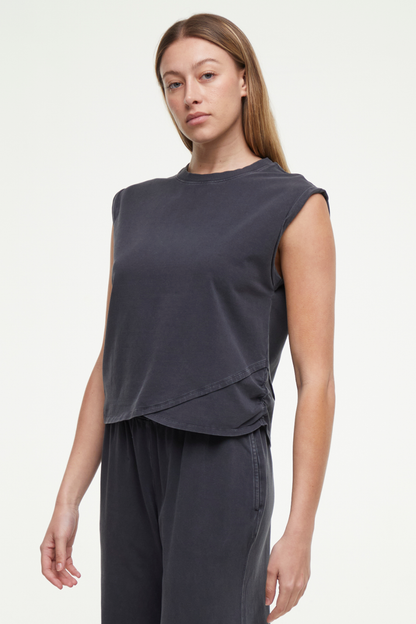 Front quarter view of woman wearing a dark gray sleeveless asymmetrical top and pants on a white background. 