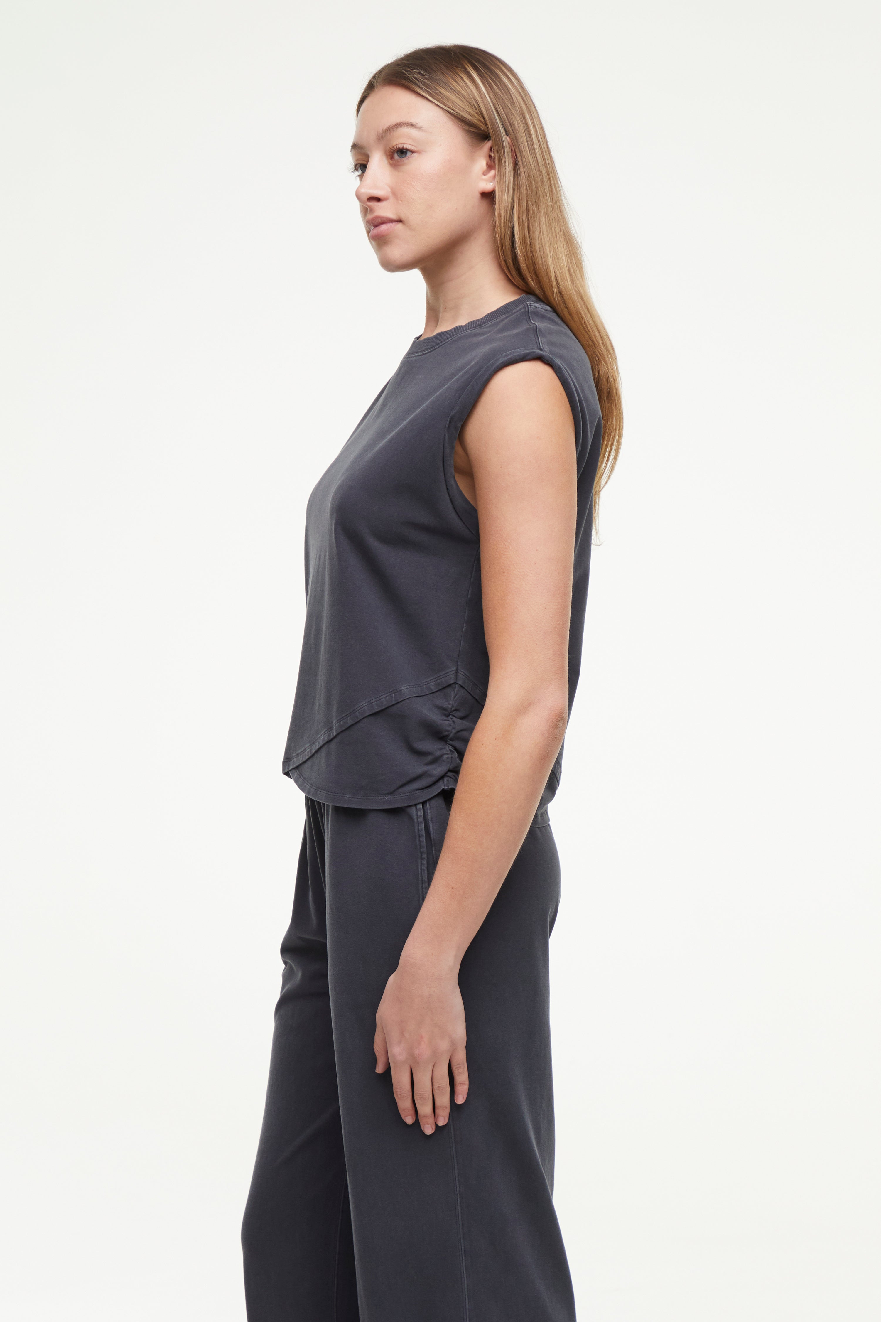 Side view of woman wearing a dark gray sleeveless asymmetrical top and pants on a white background. 