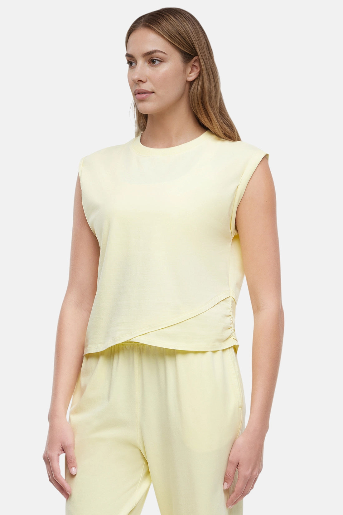 Quarter view of woman wearing a light-yellow sleeveless asymmetrical top and pants on a white background. 