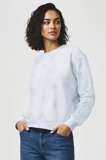 woman wearing a light blue sweatshirt with silver snowflakes pattern on it and blue jeans on a plain background