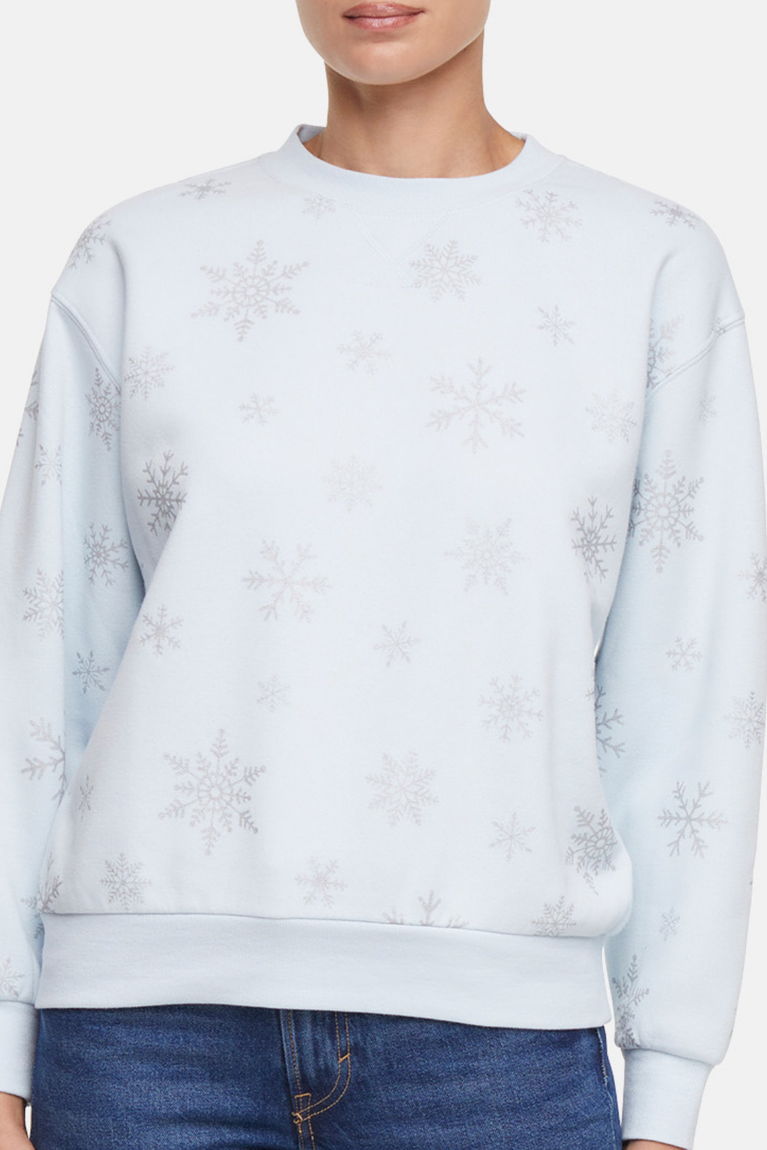 woman wearing a light blue sweatshirt with silver snowflakes pattern on it and blue jeans on a plain background
