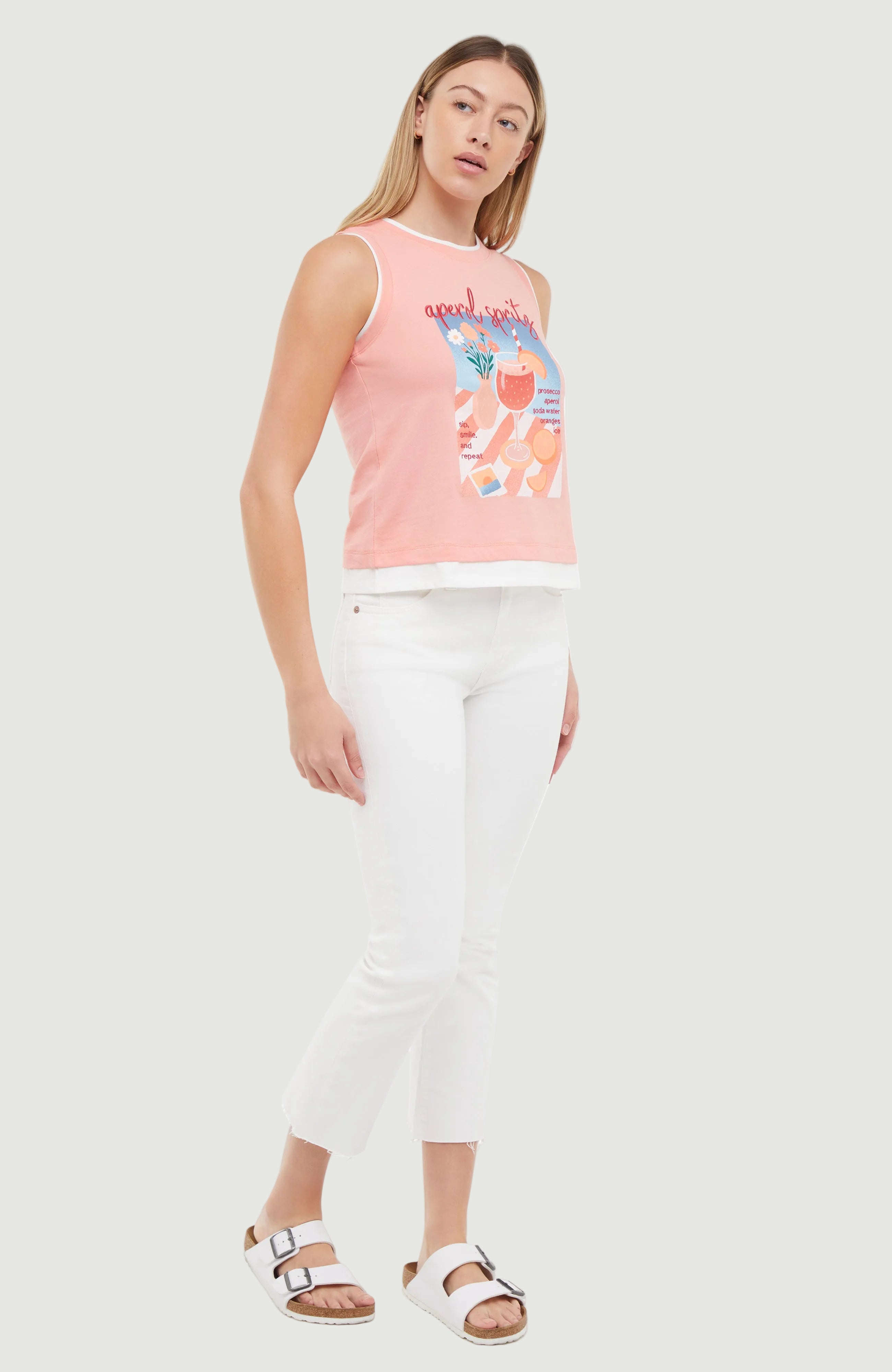 Woman wearing a pink layered sleeveless top with a graphic design and white pants on a white background