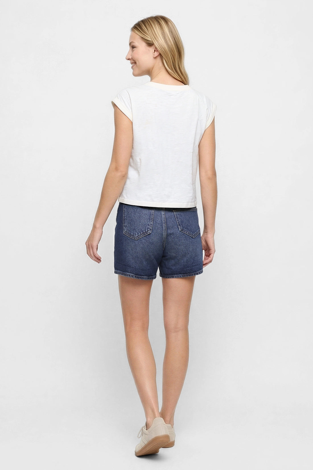 Back view of woman wearing a white cap sleeve tee and blue jeans shorts on a white background. 