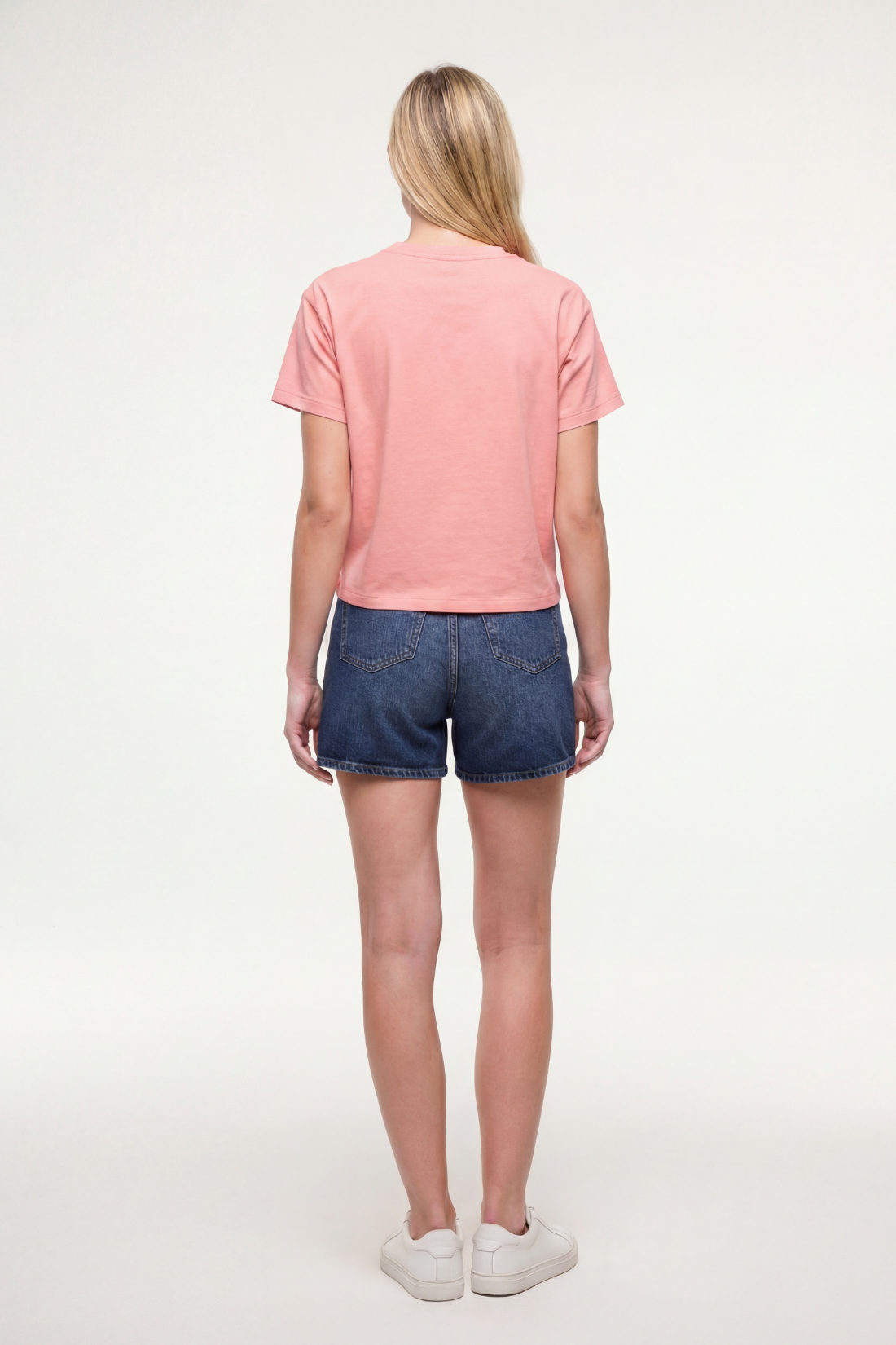 Back view of woman wearing a pale pink/red t-shirt and blue shorts on a white background.