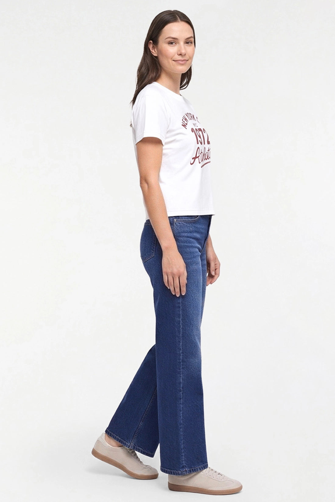 A woman modeling a white graphic t-shirt and blue flare jeans against a plain white studio background. 