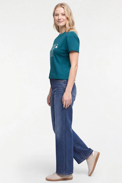 A woman modeling a teal short-sleeve graphic t-shirt and wide-leg blue jeans against a plain white studio background. 