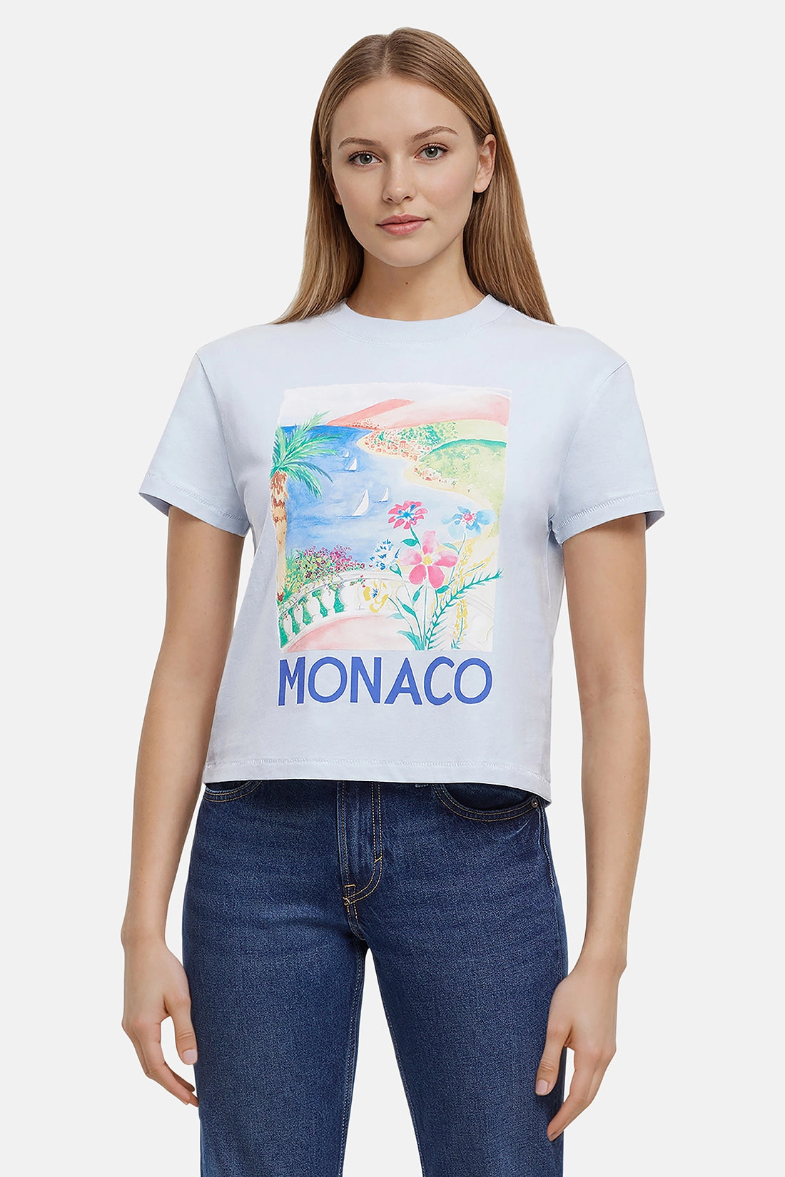 Woman wearing a light blue t-shirt with colorful beach, flowers, &amp; sailboats graphic of Monaco and the &