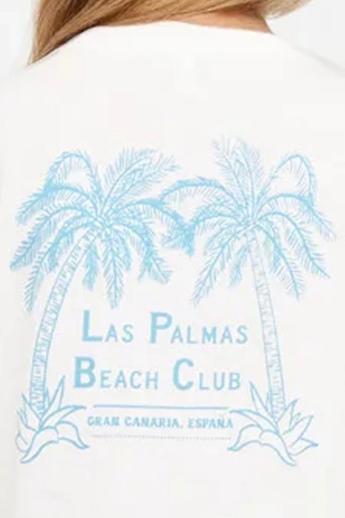 Close up back view of woman wearing a white t-shirt with "LAS PALMAS BEACH CLUB; GRAN CANARIA, ESPANA" text and graphic design featuring two palm trees on a white background. 