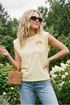 Woman wearing a yellow sleeveless shirt with a sun design, sunglasses, and holding a woven bag outdoors. 