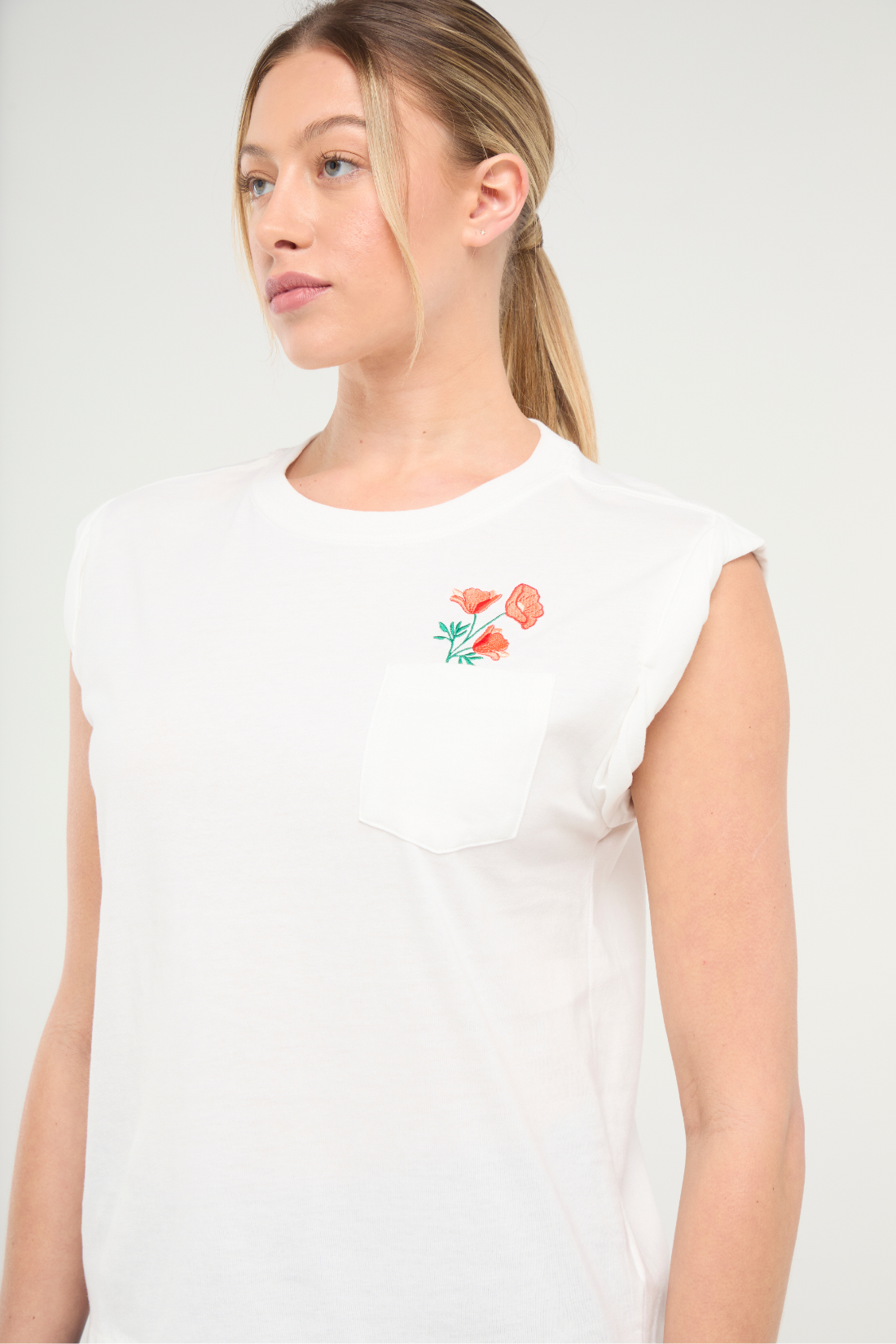 Front view of woman wearing white t-shirt with floral embroidery graphic design on top left chest pocket on a white background. 