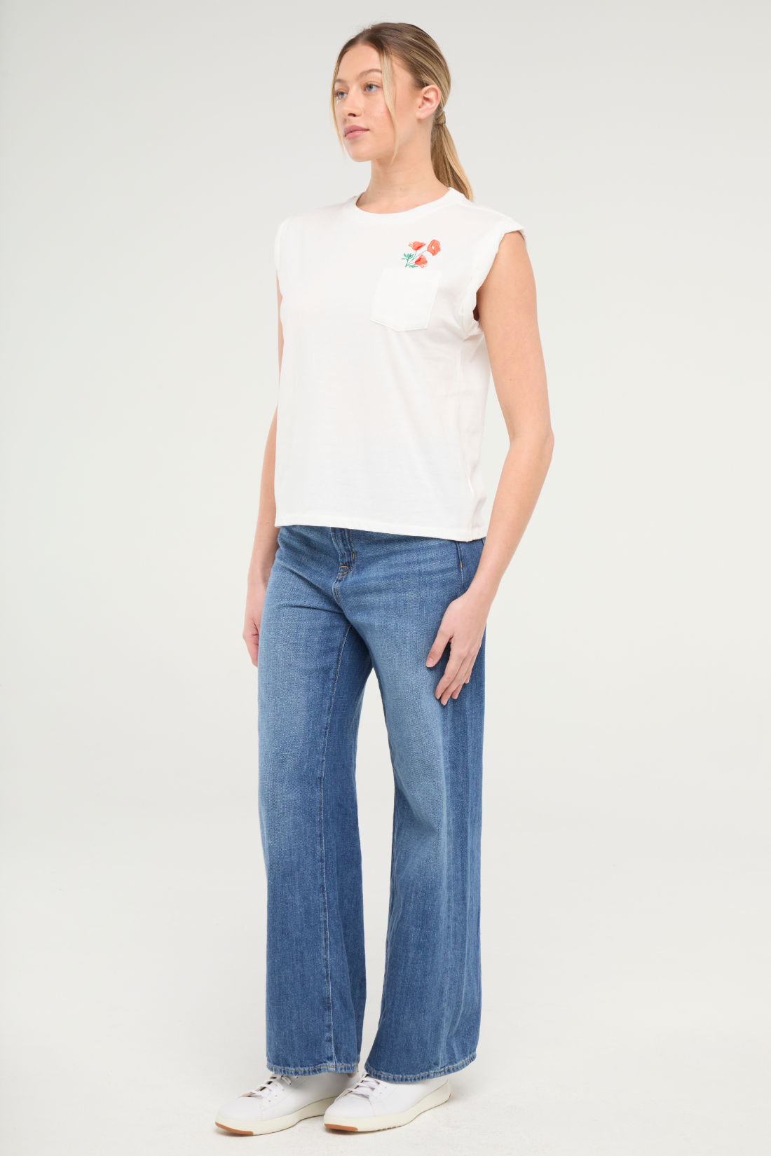 Front view of woman wearing white t-shirt with floral embroidery graphic design on top left chest pocket and blue jeans on a white background. 