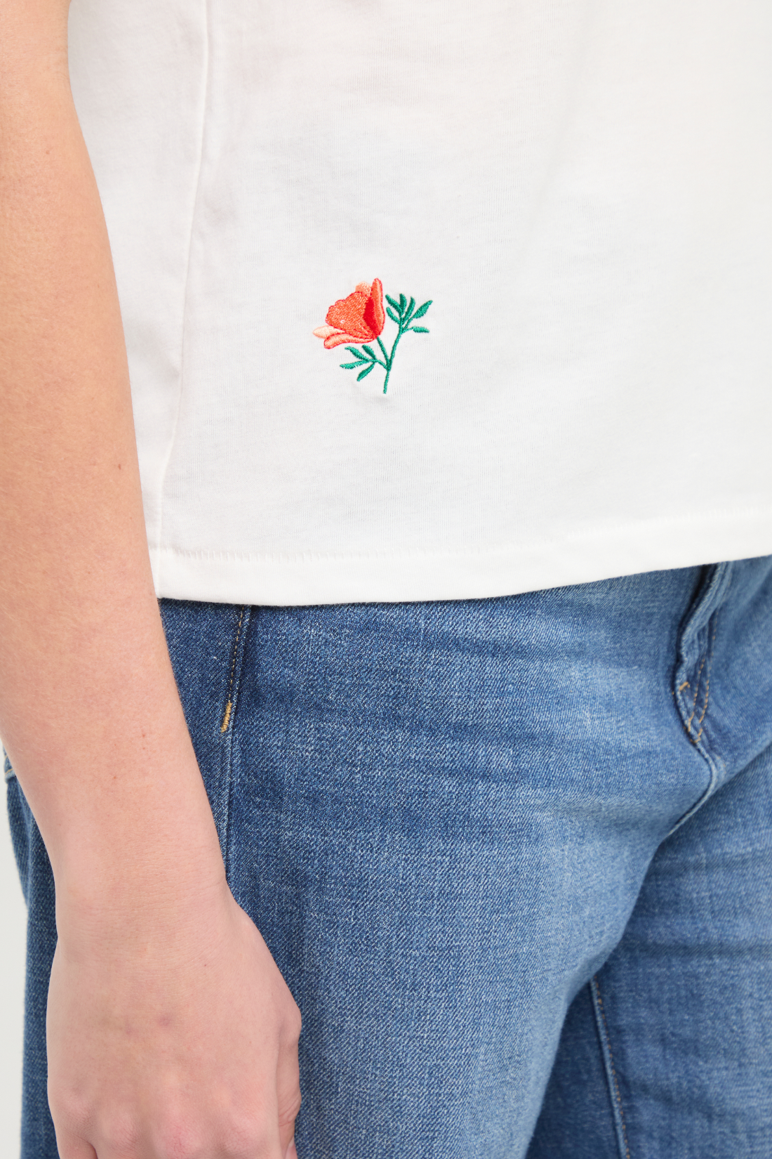 Close up front view of woman wearing white t-shirt with floral embroidery graphic design on bottom right hem and blue jeans on a white background. 