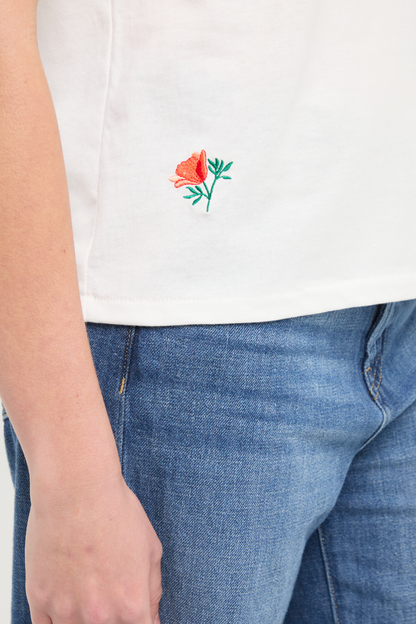 Close up front view of woman wearing white t-shirt with floral embroidery graphic design on bottom right hem and blue jeans on a white background. 