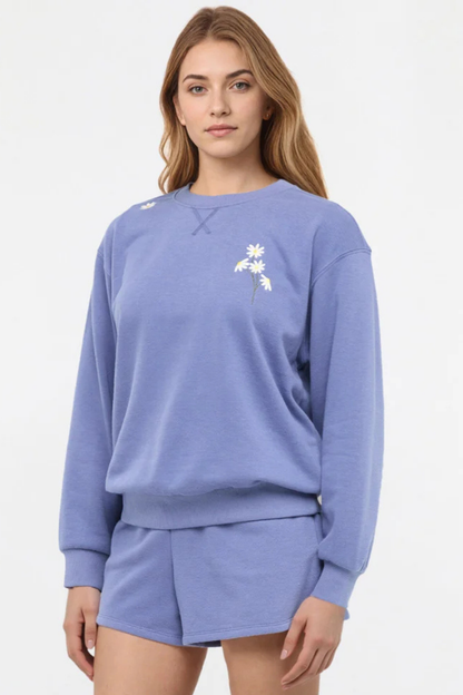 Front view of woman wearing a blue sweatshirt with floral designs on chest and shoulder and shorts on a white background. 