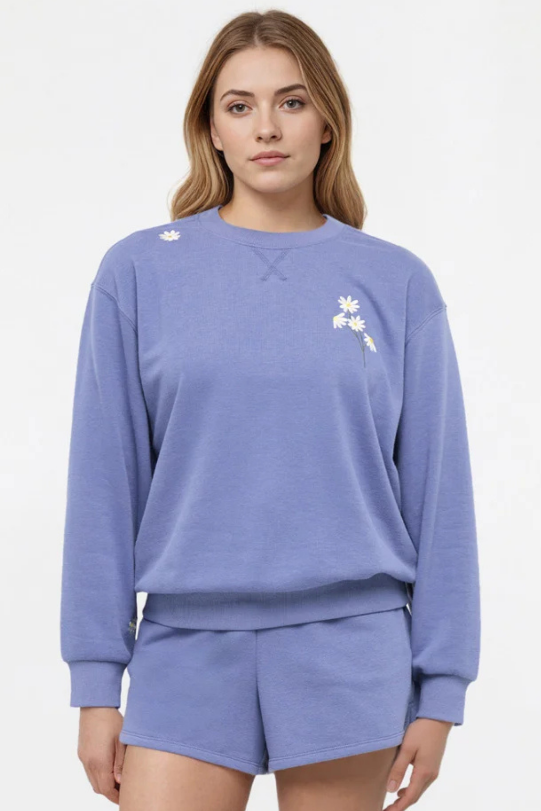 Front view of woman wearing a blue sweatshirt with floral designs on chest and shoulder and shorts on a white background.