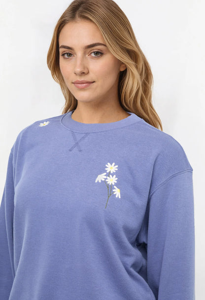 Close up view of woman wearing a blue sweatshirt with floral designs on chest and shoulder on a white background. 