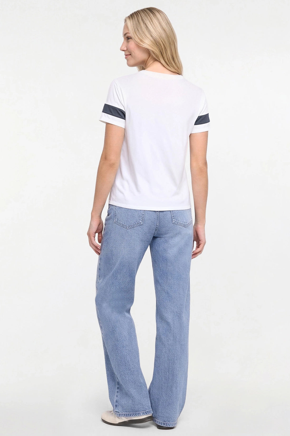 A woman is shown from the back wearing a white short-sleeve t-shirt with navy blue stripes on the sleeves and light blue wide-leg jeans, standing against a plain white studio background. 