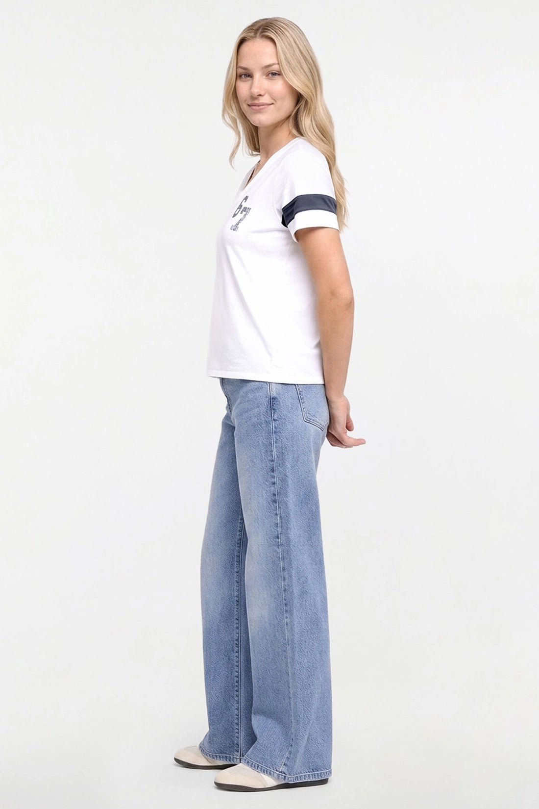 A woman modeling a white short-sleeve t-shirt with navy blue stripes on the sleeve and a small chest graphic, paired with wide-leg light blue jeans and beige shoes, against a plain white studio background. 
