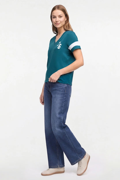 Woman modeling a teal V-neck short-sleeve t-shirt with white stripes on the sleeves and a white logo on the chest, paired with blue jeans and beige sneakers, against a plain white studio background. 