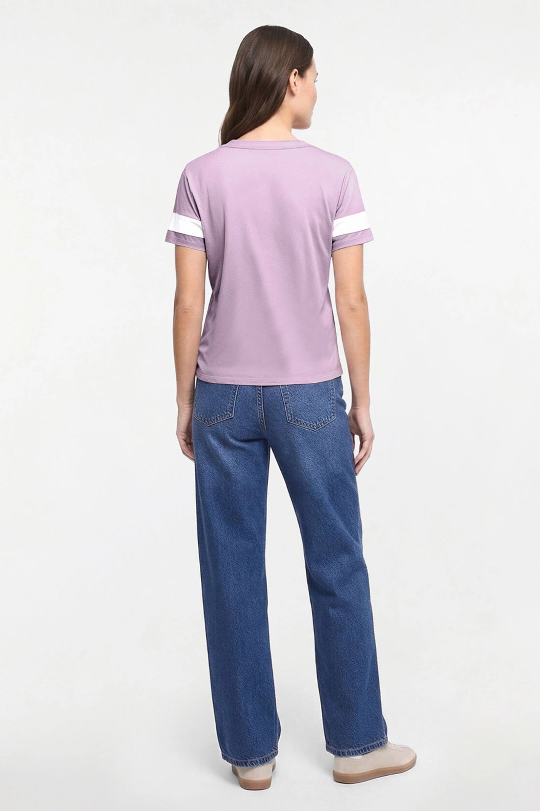 A woman viewed from the back wearing a light purple short-sleeve t-shirt with white stripes on the sleeves and blue jeans, standing against a plain white studio background.