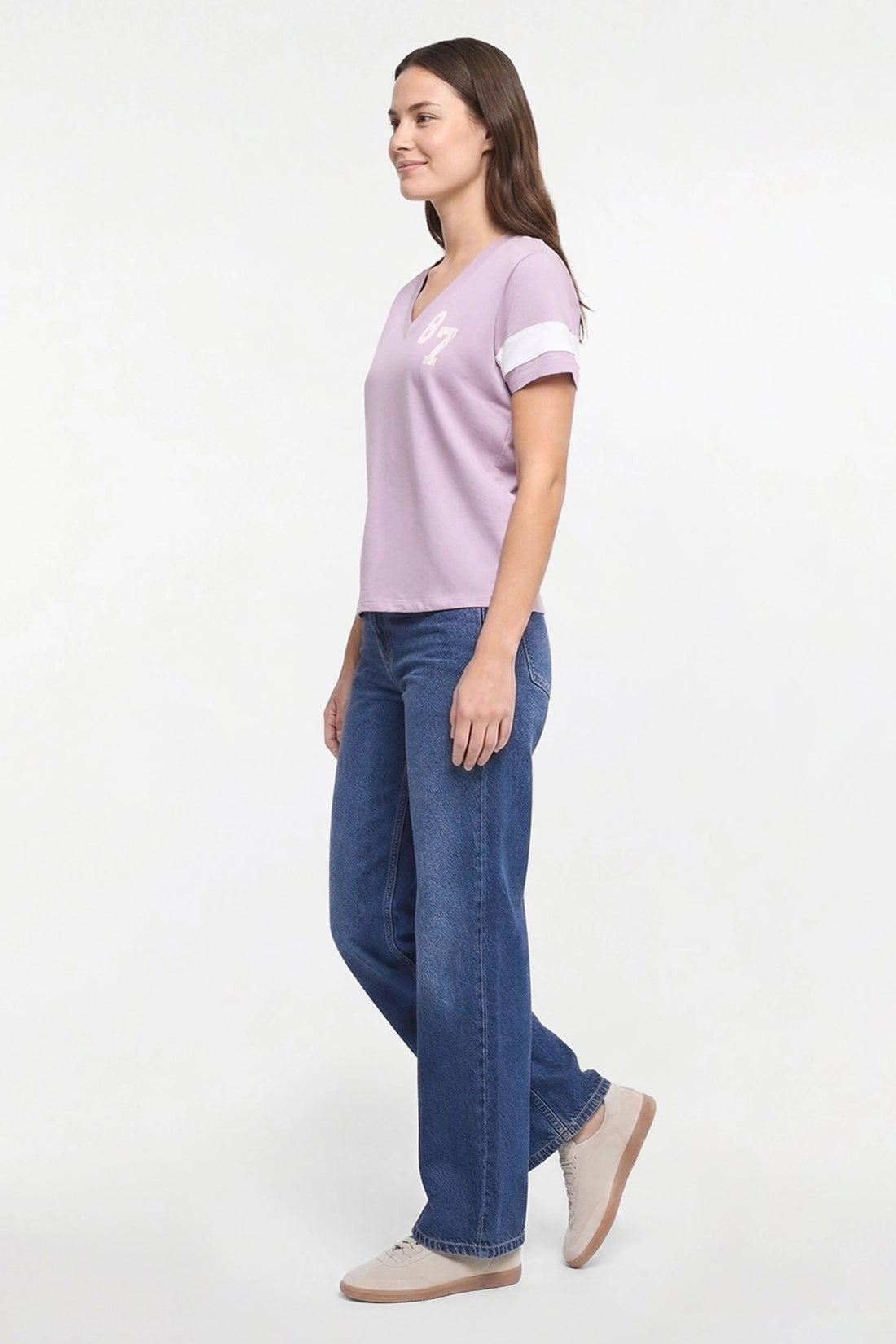 A woman modeling a light purple short-sleeve V-neck t-shirt with white sleeve stripes and a white chest graphic, paired with blue jeans and beige shoes, against a plain white studio background.