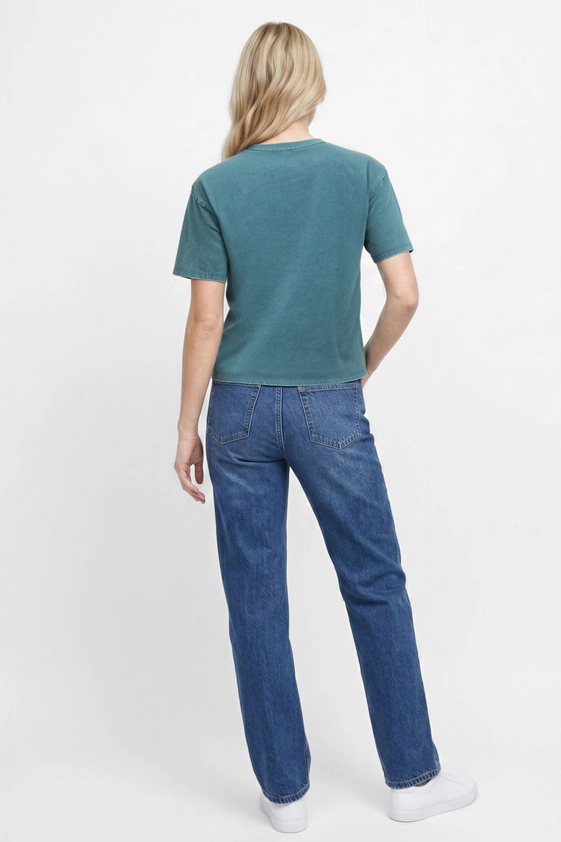 Back view of woman wearing a teal green t-shirt and blue jeans on a white background. 