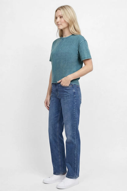 Side view of woman wearing a teal green t-shirt and blue jeans on a white background. 